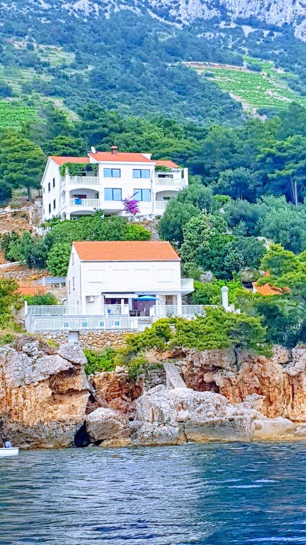 A two-story white building with a red roof, situated on a rocky coastline with a view of the sea.
