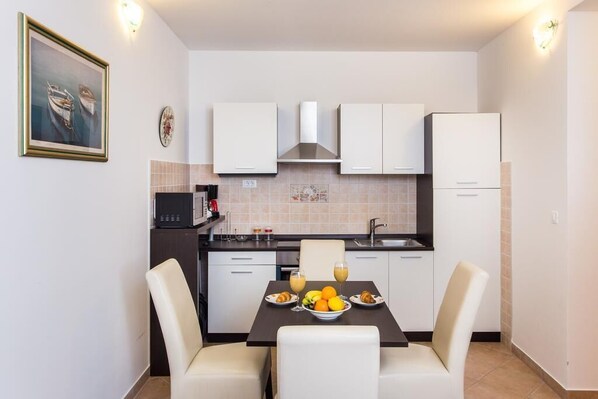 A compact kitchen with white cabinets, a black microwave, and a dining table set for two with plates of fruit and juice.