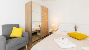 Een hotelkamer met een grijze bank, een bed met witte en gele kussens, een houten garderobe, een nachtkastje met een lamp, en een handdoek op het bed.