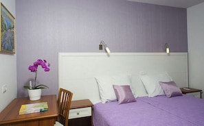 Un dormitorio con una cama, una mesita de noche, un escritorio con una silla, una planta en maceta y un cuadro en la pared.