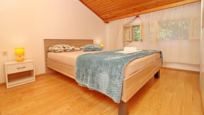 A bedroom with a wooden ceiling, a bed with a blue blanket, a nightstand with a lamp, and a window with sheer curtains.