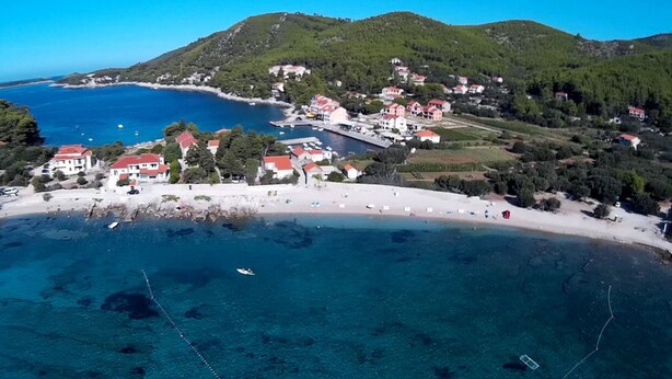 A coastal village with buildings along the beach, clear blue sea, and hilly background.