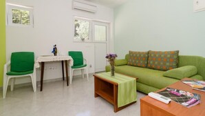 A living room with a green sofa, two green chairs, a small table with a vase of flowers, and a coffee table with a remote control and magazines.