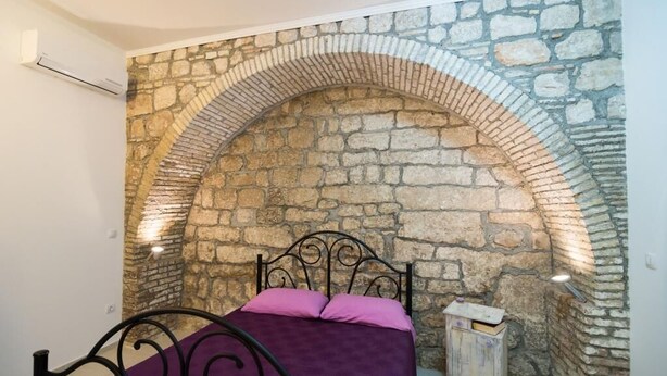 Un dormitori amb un arc de paret de pedra, un llit amb llençols porpra i un petit arca de fusta.