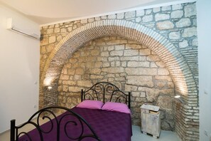 A bedroom with a stone wall arch, a bed with a purple bedspread, and a small wooden chest. - Budget Old Town - Studio with Sofa Bed - Resticeva Street (Dubrovnik)
