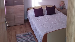 Ett sovrum med en garderob i trä, en säng med vita sängkläder och lila kuddar, ett nattduksbord med en lampa och en färgglad, vävd matta.