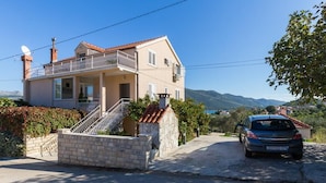 A two-story house with a balcony, a car parked in front, and a mountainous landscape in the background. - Apartments Memunić - One Bedroom Apartment with Balcony and Sea View (Ston)