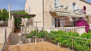 A two-story building with a tiled roof, a balcony with a white railing, and a garden area with green plants. - Milka & Miho Apartments - Double or Twin Room 1 (Second Floor) (Dubrovnik)