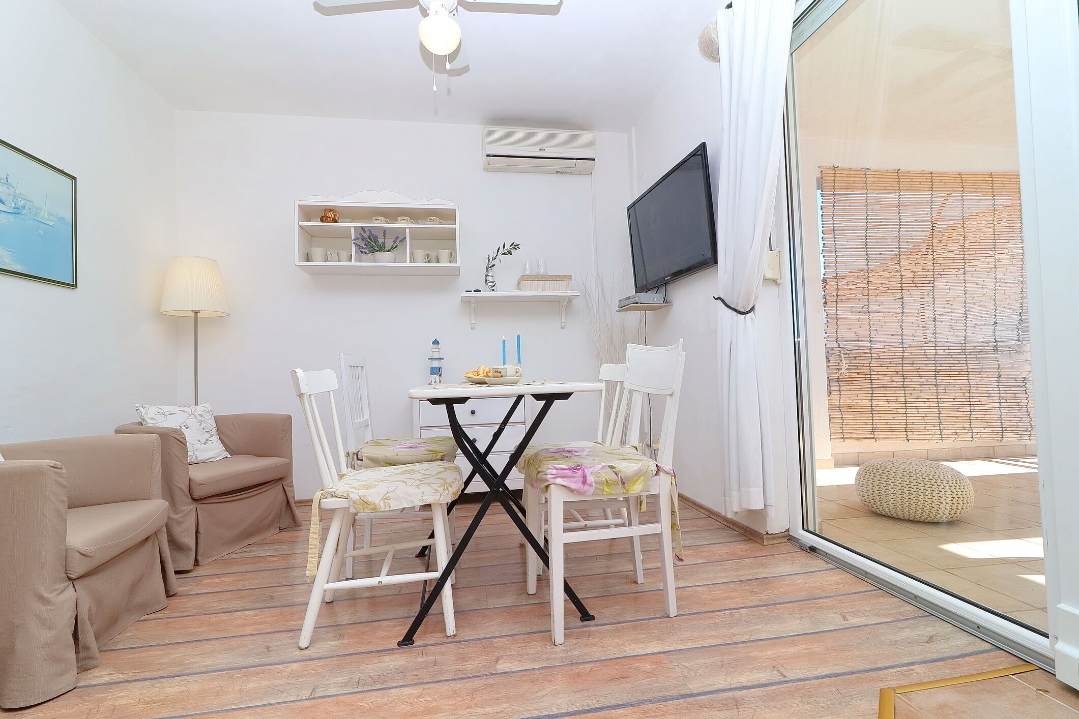 A compact living space with a sofa, a dining table with chairs, a TV mounted on the wall, and a large window with blinds.