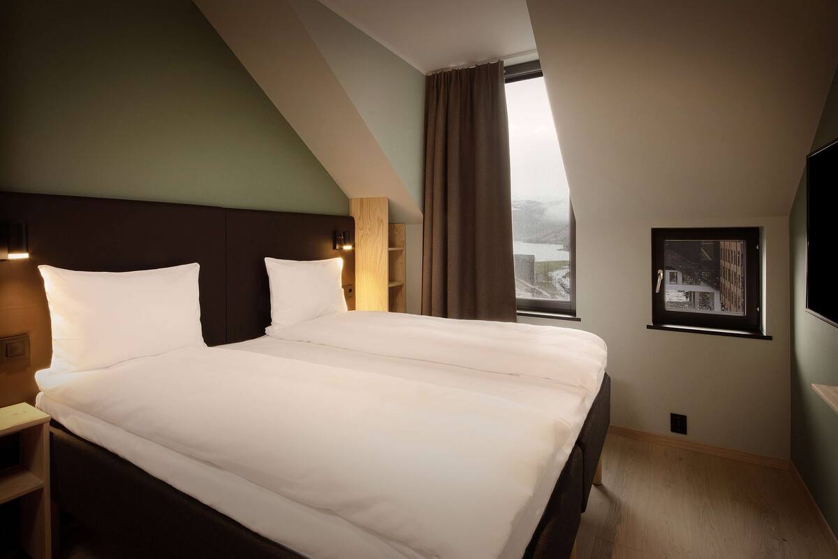 A hotel room with a large bed, a nightstand, a wall-mounted TV, and a window with a view of the city.