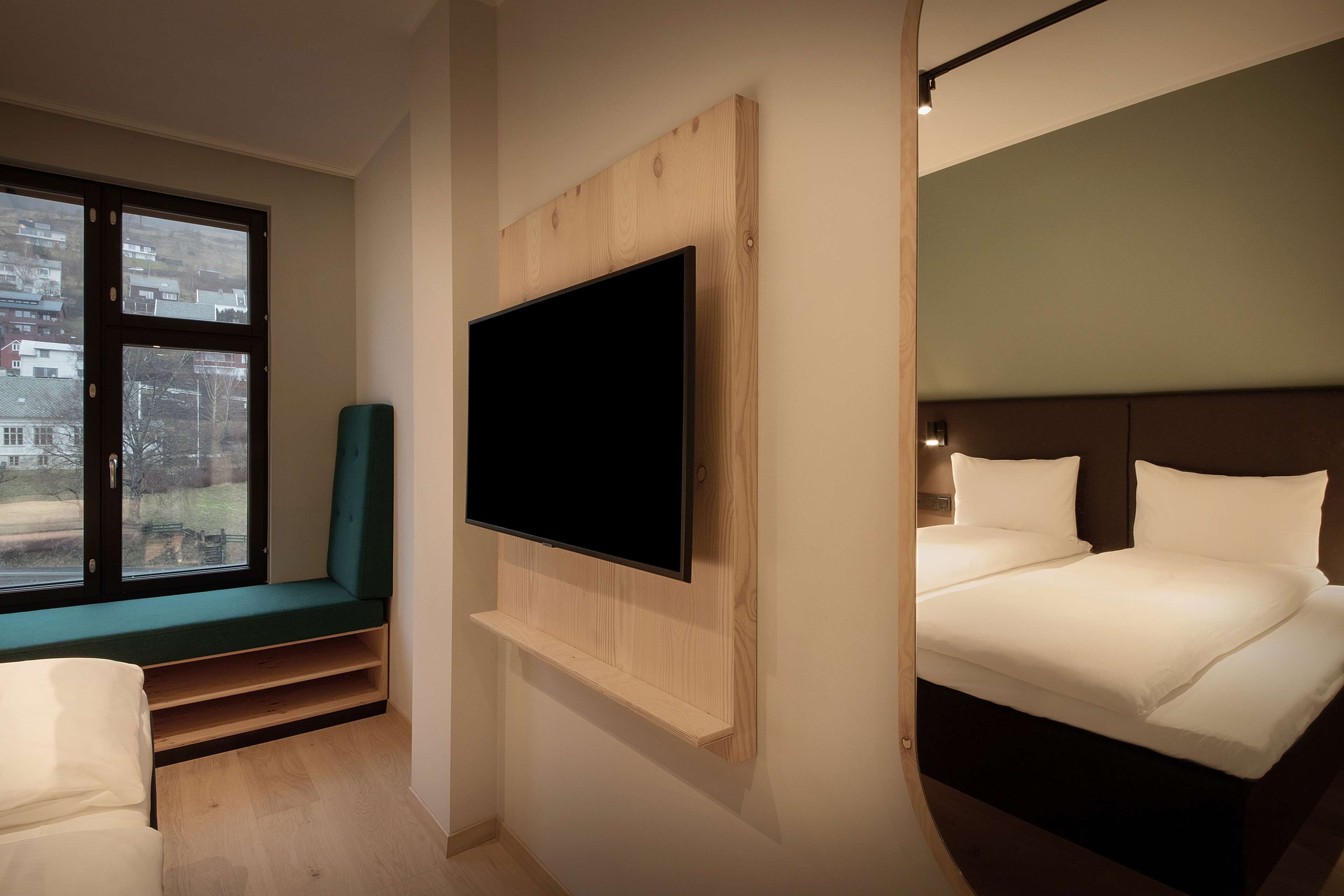 Quarto de hotel moderno com uma televisão grande fixada em um painel de madeira, uma cama com roupa branca, um banco com almofada verde e uma janela com vista para a cidade.