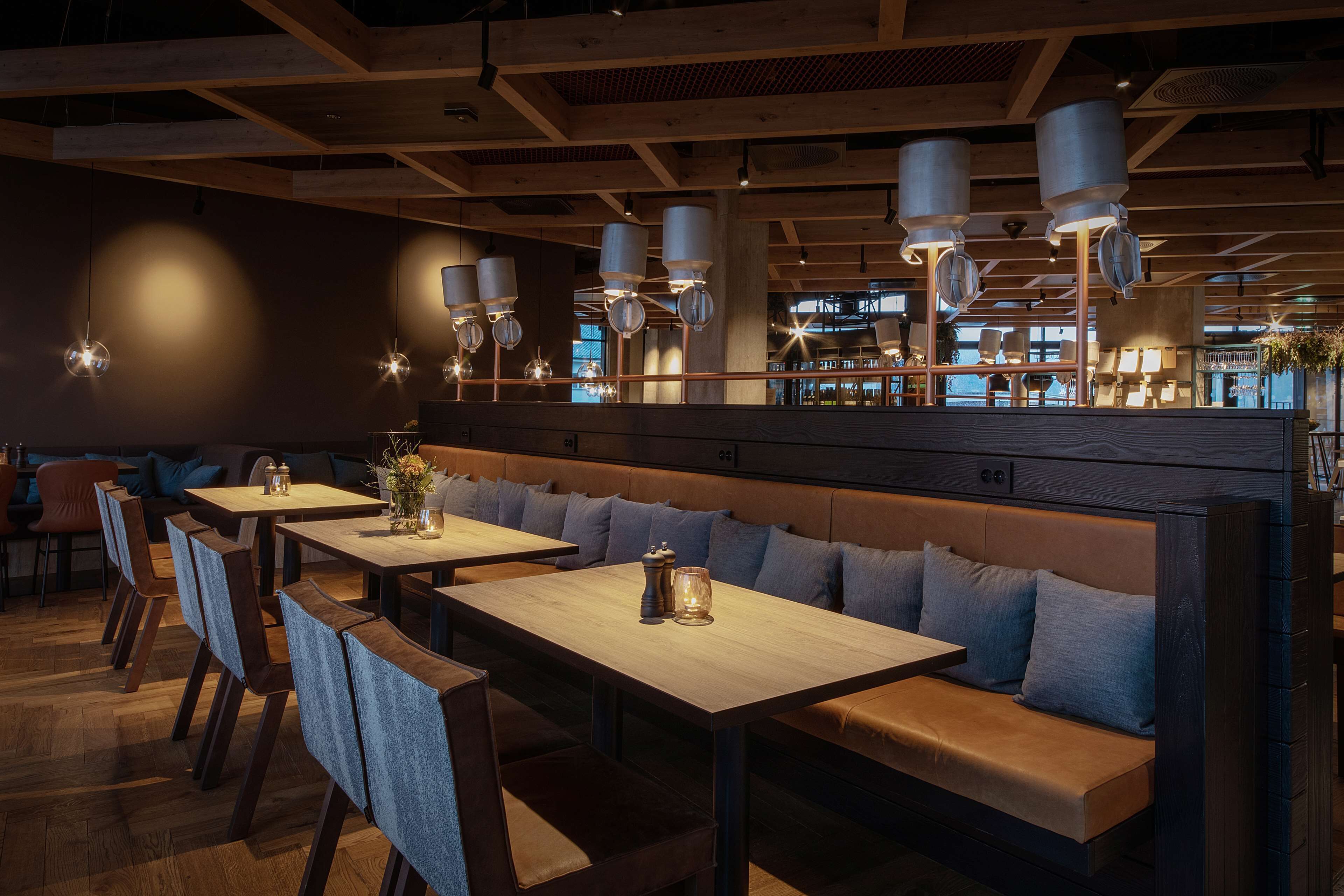 Interior de restaurante con mesas de madera, bancos acolchados, lámparas colgantes y vista al área de la cocina.
