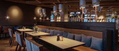 Interior de restaurante con mesas de madera, bancos acolchados, lámparas colgantes y vista al área de la cocina.