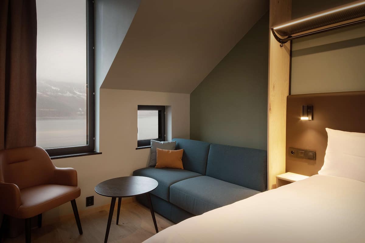 A modern hotel room with a bed, a sofa, a small table, and a chair. There is a window with a view of mountains.