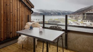 A wooden balcony with a table and chairs, overlooking a snowy landscape with mountains and a bridge.