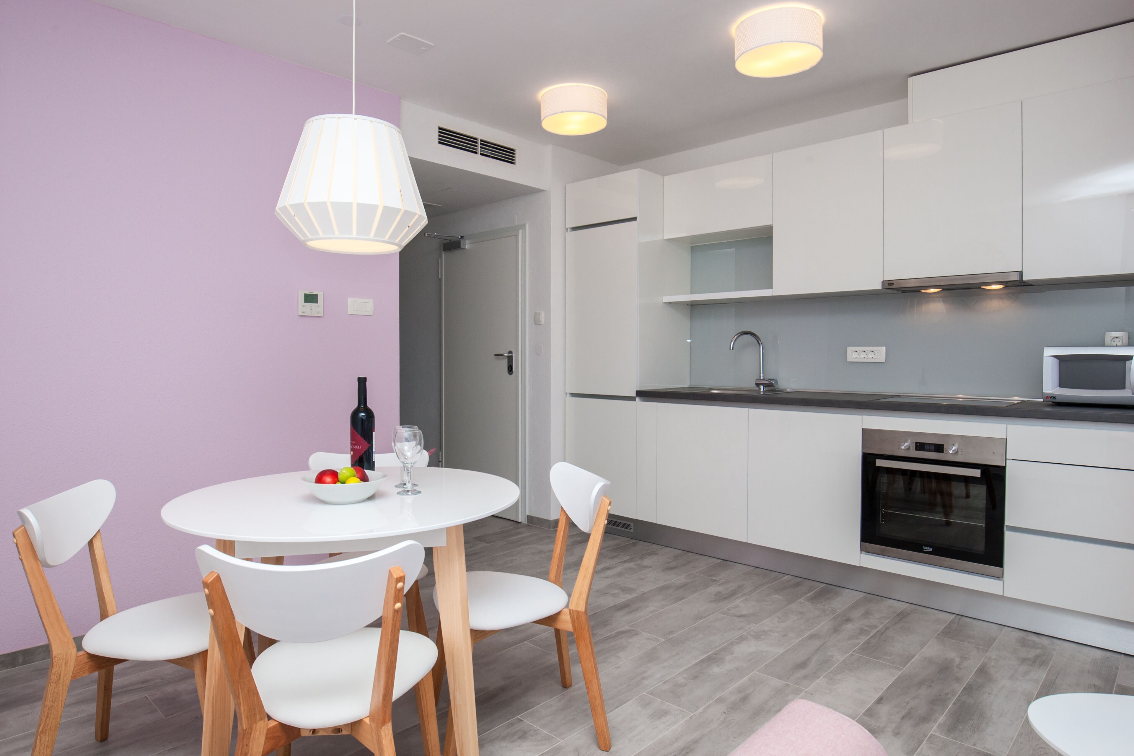 Une cuisine moderne avec des armoires blanches, un four encastré et un micro-ondes. On y trouve un coin repas avec une table et des chaises blanches, et un luminaire suspendu au-dessus de la table.