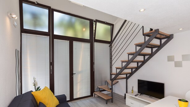A modern living room with a dark sofa, yellow cushions, a white table, a TV, and a wooden staircase. - Apartments Pomet - Premium Duplex Studio with City View (Puhiera) (Dubrovnik)
