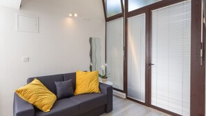 A modern living room with a dark grey sofa, yellow cushions, a small white table with a plant, and a large sliding glass door with blinds. - Apartments Pomet - Premium Duplex Studio with City View (Puhiera) (Dubrovnik)