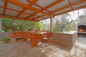 Relax in a serene outdoor space with a wooden table, stone bench, and fireplace under a covered pergola amidst lush greenery.