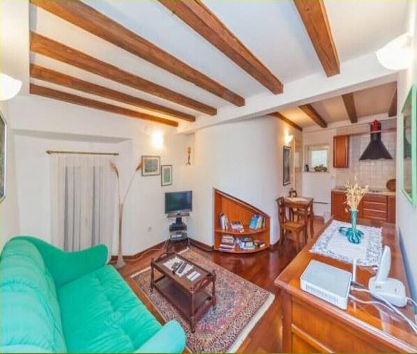 A cozy living room with a green sofa, wooden beams on the ceiling, a bookshelf, and a dining area with a table and chairs.