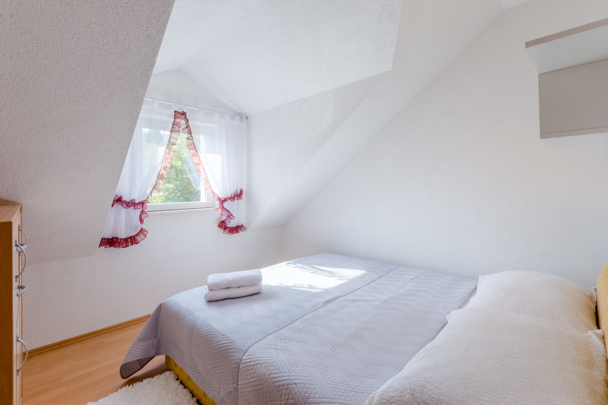 A bedroom with a sloped ceiling, a bed with white bedding, a window with red curtains, and a wooden floor.