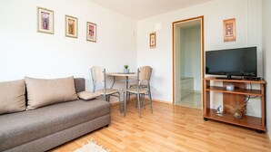 A living room with a sofa, a small table with chairs, a TV on a wooden stand, and framed pictures on the wall.