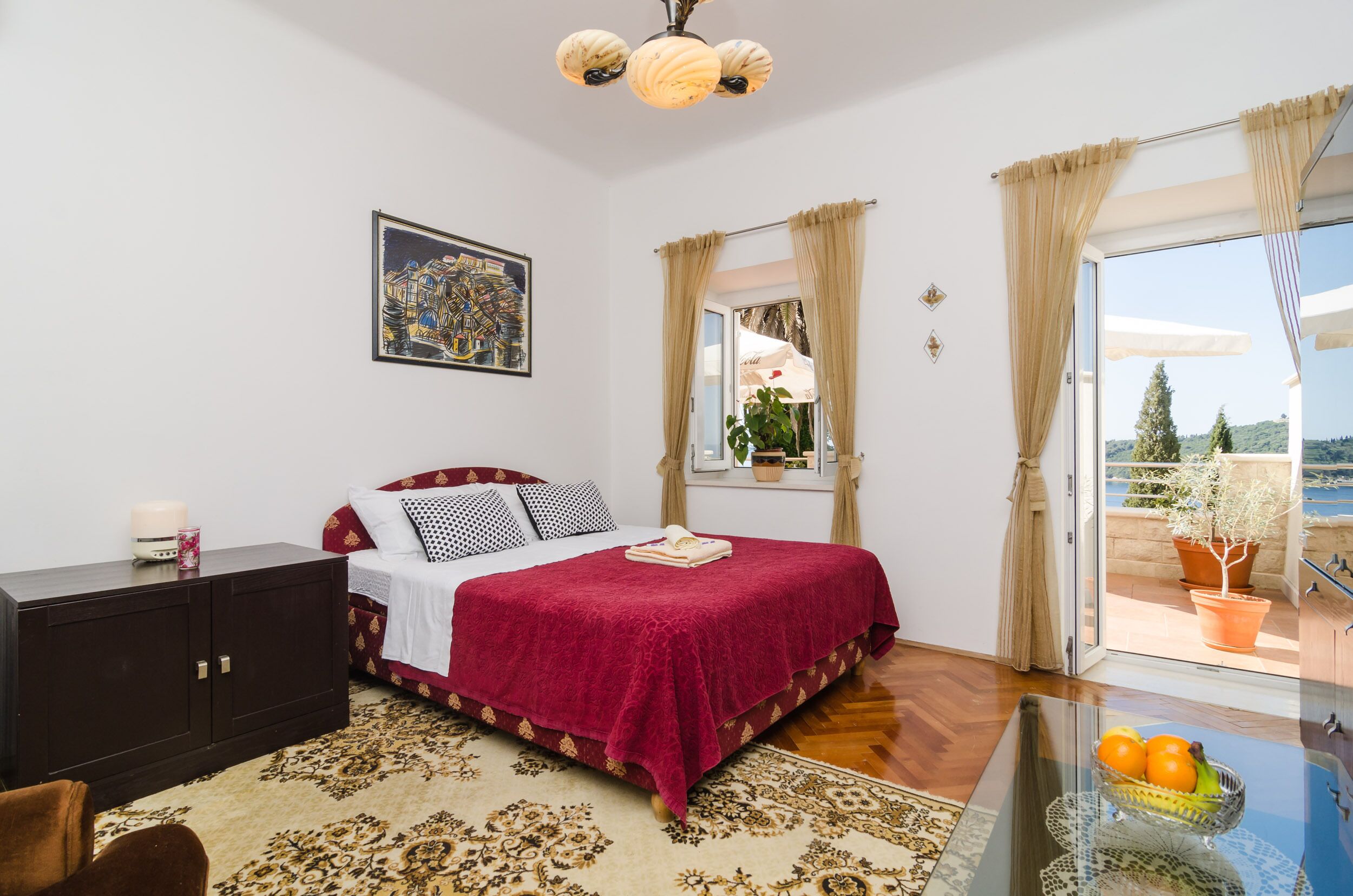 A bedroom with a bed, a dresser, a window with curtains, a balcony with potted plants, and a view of the sea.