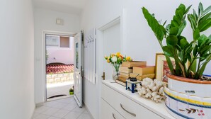 A hallway with a sliding door leading to an outdoor area, a white wall with a mirror, and a shelf with a potted plant, books, and decorative items. - Guesthouse Matusic - Double Room (Dubrovnik)