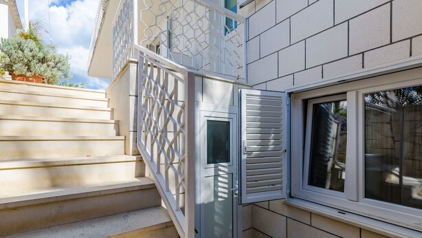 Un coin d’un bâtiment avec une porte blanche, une fenêtre avec des stores et un escalier menant à l’étage supérieur.