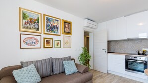 Uma sala de estar moderna com um sofá marrom, uma pequena planta e uma cozinha com armários brancos e um forno.