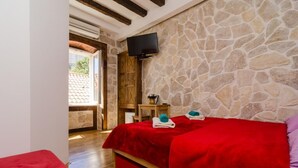 Una habitación con pared de piedra, techo de madera, cama con ropa de cama roja, televisión montada en la pared y una pequeña mesa de madera con un florero.