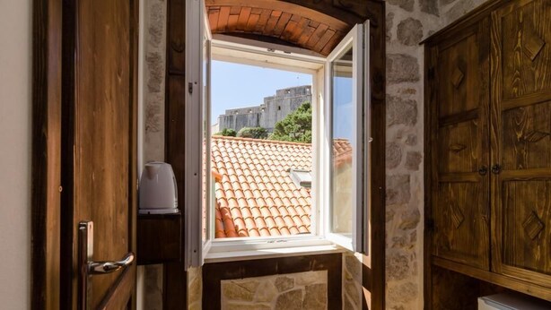 Una habitación con pared de piedra, puerta de madera, ventana que ofrece vista a un tejado y un edificio, y un armario de madera.