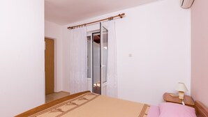 Ein Schlafzimmer mit einem Bett, einem Nachttisch mit Lampe, einem Fenster mit Vorhängen und einer Tür zu einem anderen Raum.