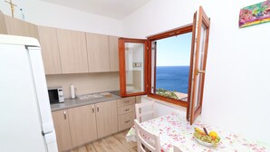 Eine kleine Küche mit Holzausstattung, einem weißen Kühlschrank, einer Mikrowelle und einem Tisch mit einem blumigen Tischläufer. Ein Fenster mit offenem Holzrahmen bietet Blick auf das Meer.