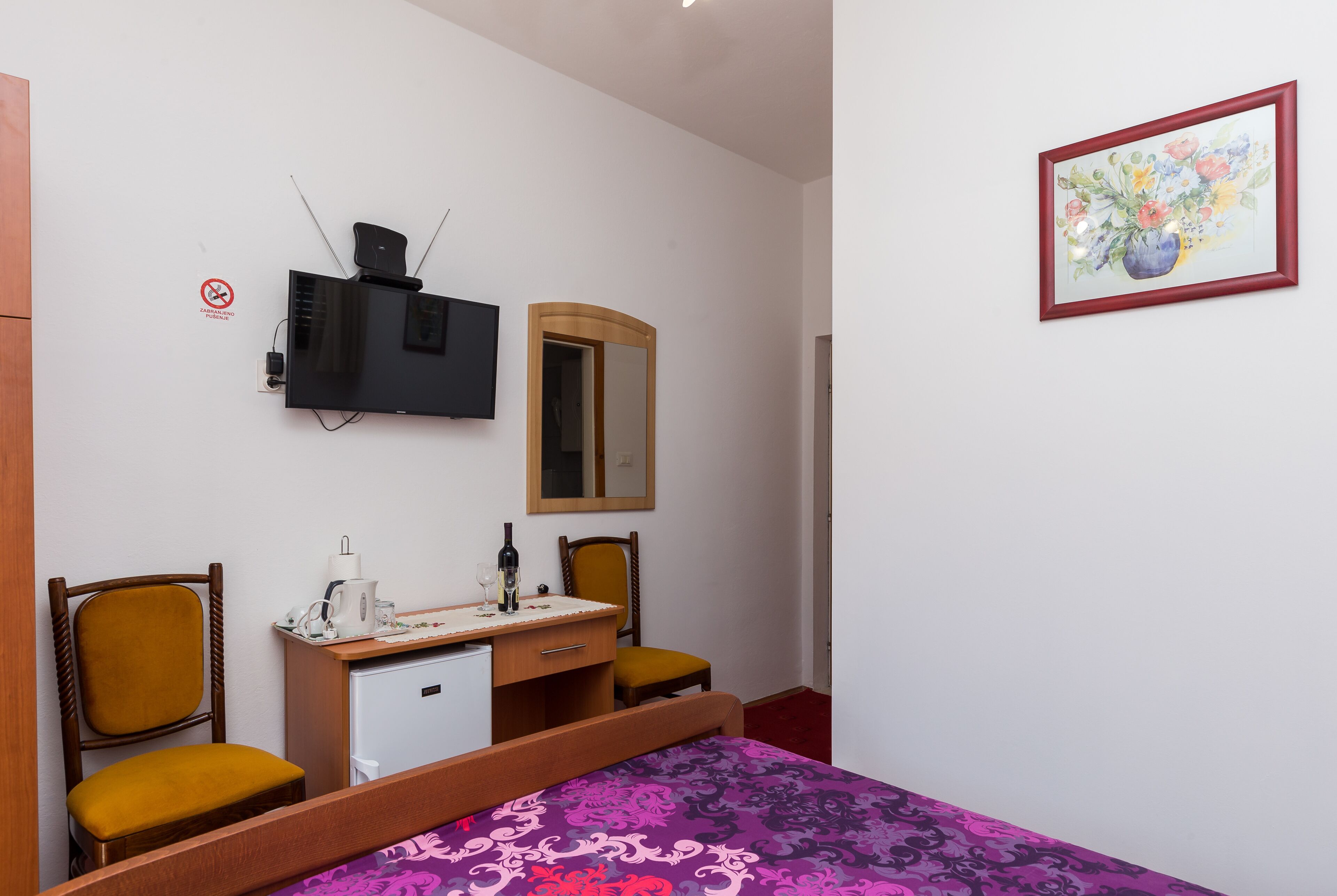 A bedroom with a bed, a desk with a mirror, a television, and a painting on the wall.