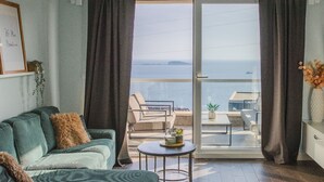 A living room with a sofa, a small round table, and a view of the sea through a sliding glass door.