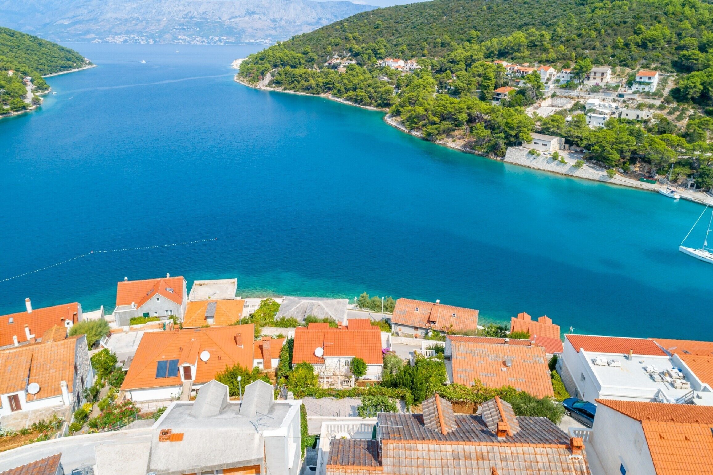 A coastal village with terracotta-roofed buildings, a marina, and a clear blue sea.