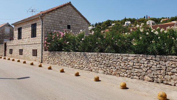 A stone building with a tiled roof, a stone wall with bollards, and flowering bushes.