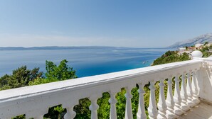 Une vue sur la côte, prise depuis un balcon doté d’une balustrade blanche, donnant sur une mer bleue et limpide et sur des montagnes lointaines.