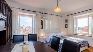 Ein Schlafzimmer mit Bett, Nachttischen, einem Tisch mit Stühlen und Blick auf die Stadt durch große Fenster.