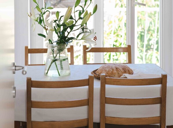 Une table à manger en bois recouverte d’une nappe blanche, deux chaises en bois, un vase avec des fleurs blanches et une miche de pain posée sur la table.