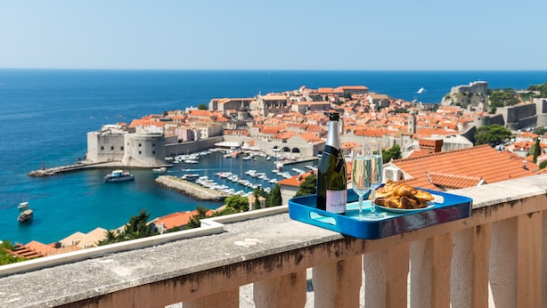 A balcony with a tray containing a bottle of wine, a glass, and pastries, overlooking a coastal town with historic buildings and a marina. - Apartments Bonavista - Superior Two Bedroom Apartment with Balcony and Sea View (A1) (Dubrovnik)