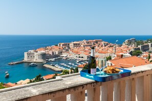 A balcony with a tray containing a bottle of wine, a glass, and pastries, overlooking a coastal town with historic buildings and a marina.
