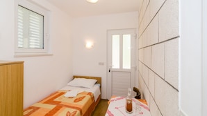 A small, clean room with a bed, a table with a bottle and glass, a window with blinds, and a door with a handle. - Katarina rooms - twin room (Dubrovnik)
