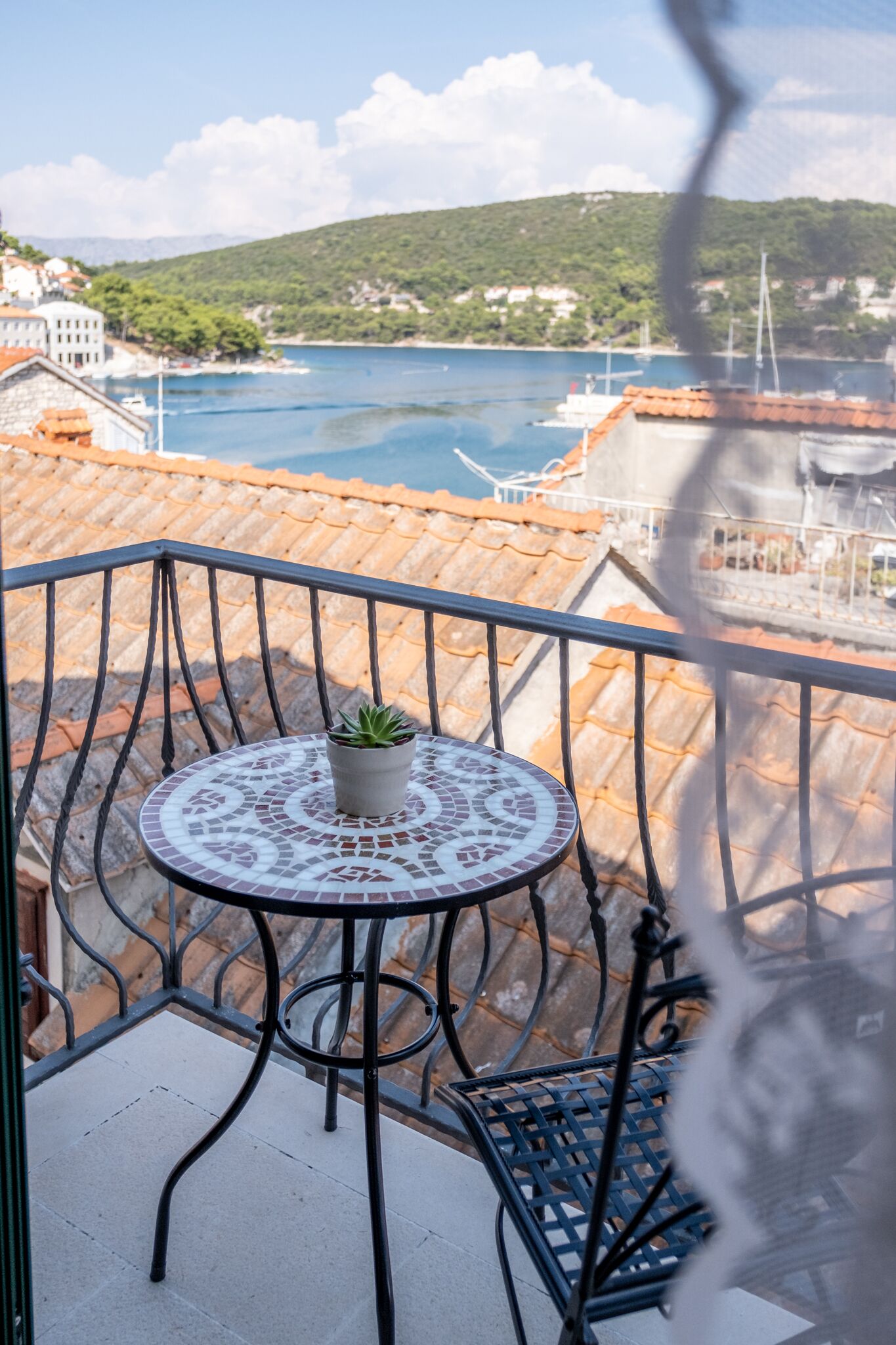 A balcony with a small table and a potted plant, overlooking a body of water and a hilly landscape.