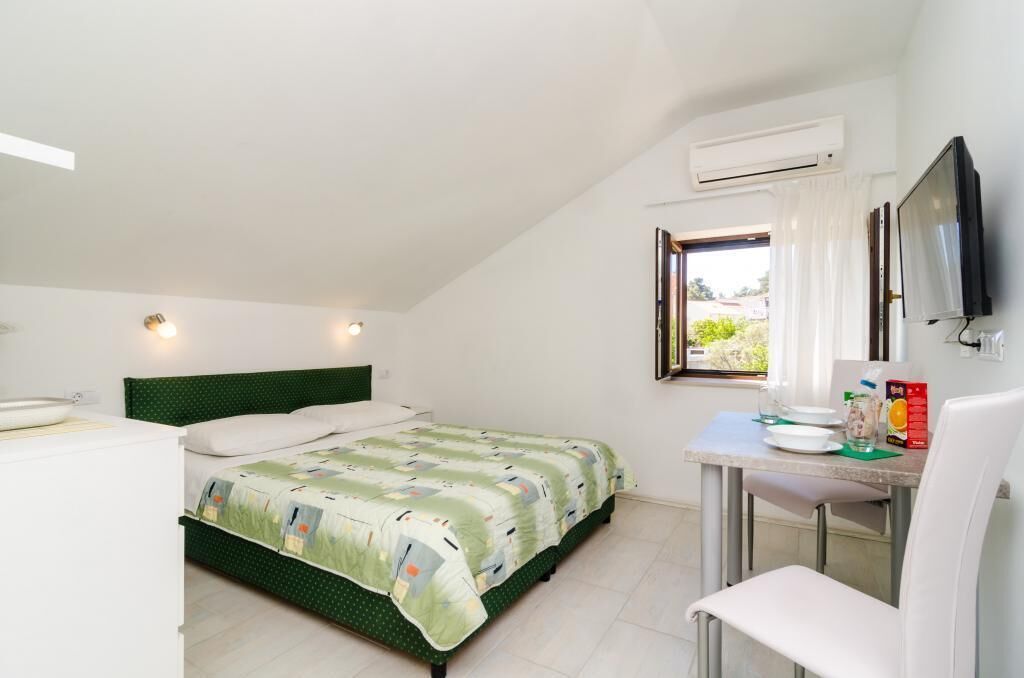 Een kleine, nette kamer met een bed, een bureau en een stoel. Er is een raam met gordijnen, een airconditioning en een flatscreen televisie aan de muur bevestigd.