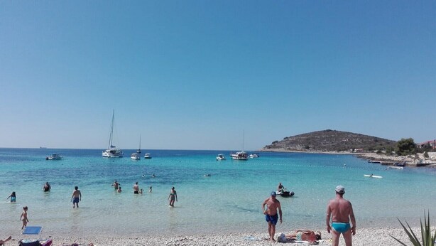Una playa con agua turquesa cristalina, gente nadando y caminando, barcos anclados a lo lejos, y una colina rocosa al fondo.