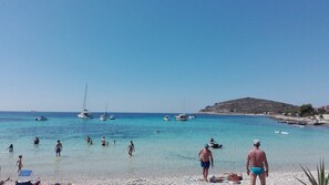 A beach with clear turquoise water, people swimming and walking, boats anchored in the distance, and a rocky hill in the background.