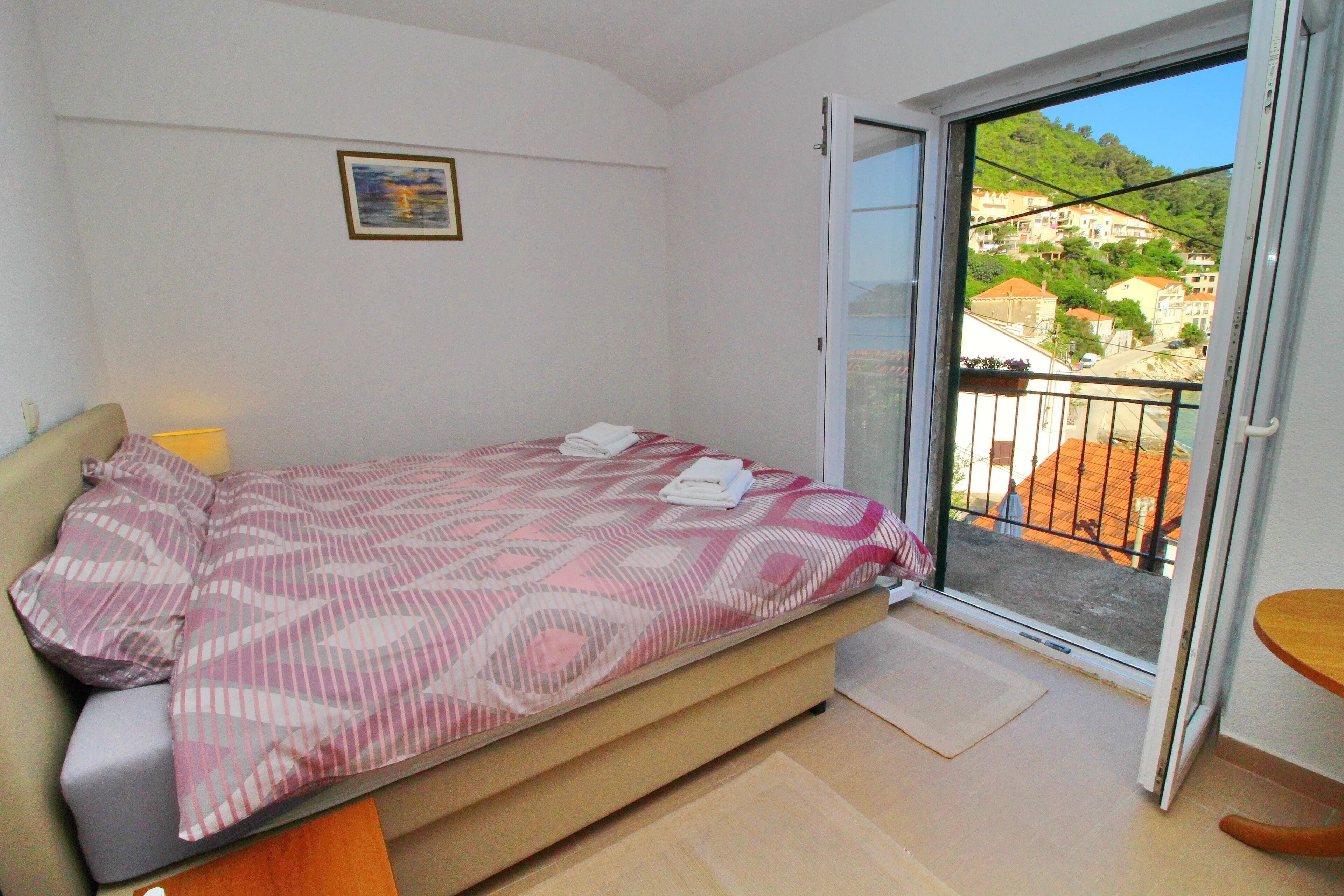 Une chambre avec un lit, une petite table, une chaise, une porte donnant sur un balcon et une vue sur des immeubles et de la verdure.
