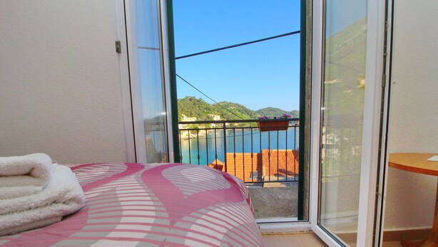 A bedroom with a bed, a window, and a view of a balcony with a railing and a wooden chair. - Apartment Mambo , Mljet - Duplex Apartment with Terrace and Sea View (Sobra)